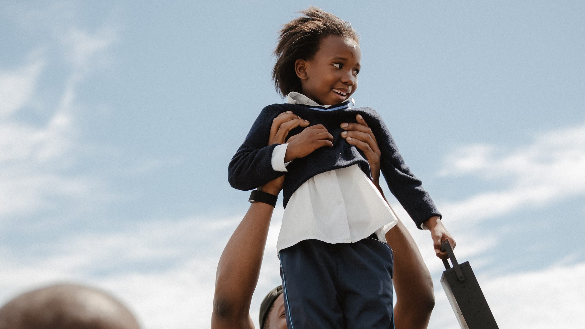 A child lifted high above a crowd of schoolchildren, laughing, against a blue sky.