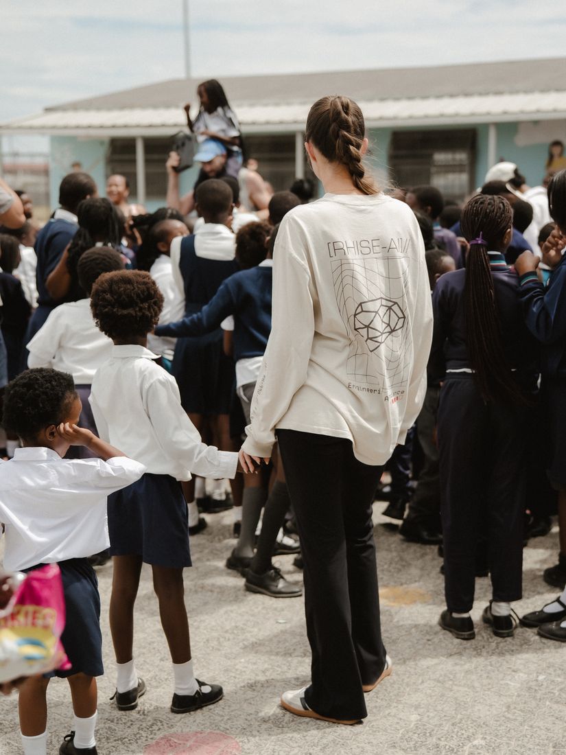 A visitor in a RHISE AI shirt holding a child’s hand in the school yard.