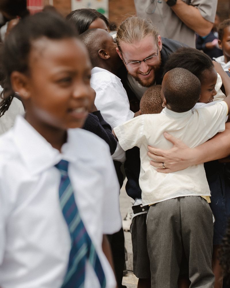 A visitor kneeling to hug two children in school uniform.