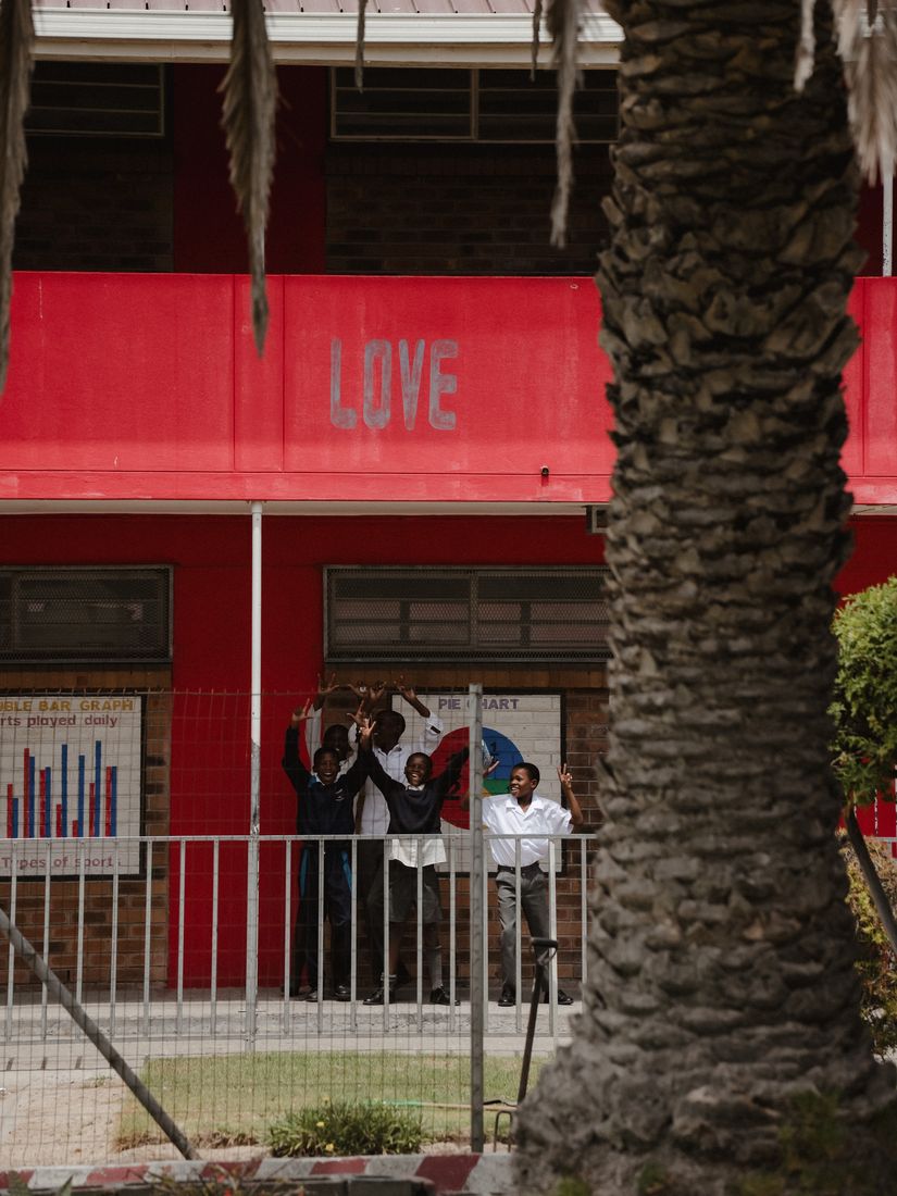 Children waving from a balcony beneath the word LOVE painted on a red school wall.